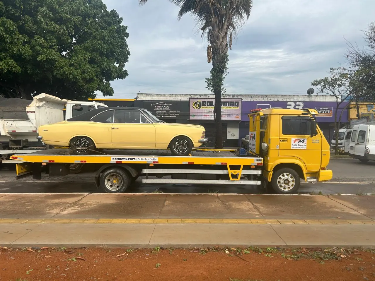 Chevrolet Opala clássico amarelo com teto preto sobre o guincho plataforma da FM Auto Socorro, parado junto ao canteiro de uma avenida
