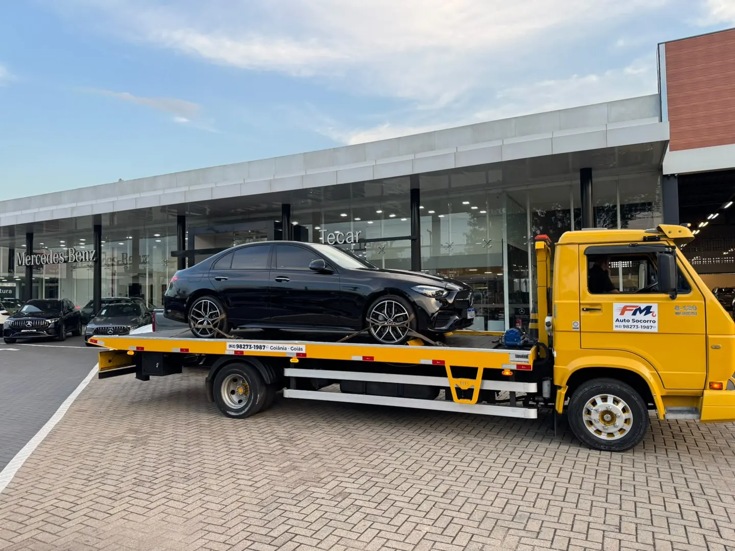 Caminhão guincho plataforma amarelo da FM Auto Socorro com um sedã Mercedes-Benz preto amarrado na carroceria, em frente a uma concessionária Mercedes-Benz